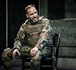 Macbeth in battle uniform and holding glass of booz sits on a chair in front of the steps of the bunker